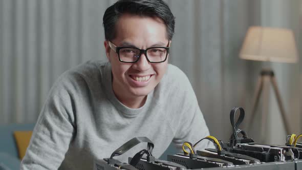 Close Up Of Asian Man Smiling To Camera While Using Laptop And Mining Rig For Mining Cryptocurrency