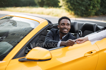  Service. Test Drive Concept. Young African American man sitting inside new yellow cabriolet car while taking keys from female seller.