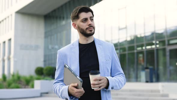 Successful Bearded Man With Tablet and Coffee Cup Walking Between Business Buildings and Smiling