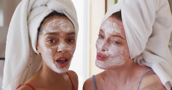 Diverse happy female friends wearing towels on heads and cleansing masks taking selfie at home