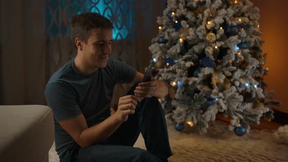 Young Caucasian Man Using Phone Sitting Near Cozy Christmas Tree
