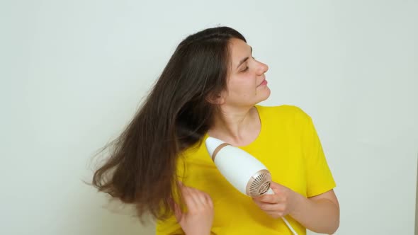 Brunette Woman Dries Her Long Hair with a Hair Dryer and Smiles on a White Background