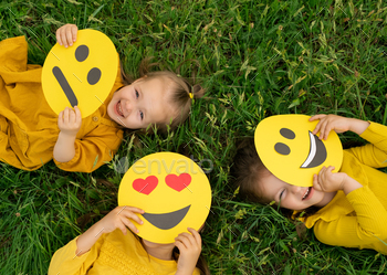 ind the faces of emoticons with different emotions : happy, sad and in love. Top view. Children are lying on the grass looking up. Emoji smile day