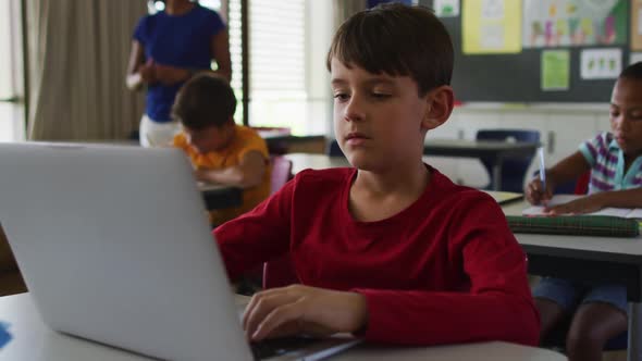 Portrait of happy caucasian schoolboy sitting at classroom, using laptop, looking at camera