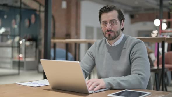 Young Man Smiling at Camera While Using Laptop in Office