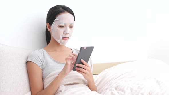 Woman apply facial mask and using cellphone on bed 