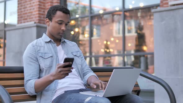 African Man Using Smartphone and Laptop Sitting on Bench
