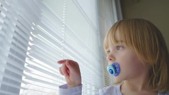 A little girl with a pacifier in her mouth looks out a window and plays with the blinds