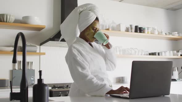 Happy african american woman with beauty mask on face, drinking coffee and using laptop in kitchen