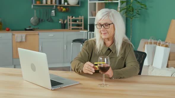 Happy Elderly Woman with a Glass of Wine Shopping Online From Home