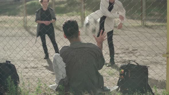 Back View of African American Bullied Man Sitting at Mesh Fence As Caucasian Guys Throwing Ball at