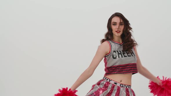 Young Cheerleader with Red Pompoms in Uniform is Dancing on White Background in Studio