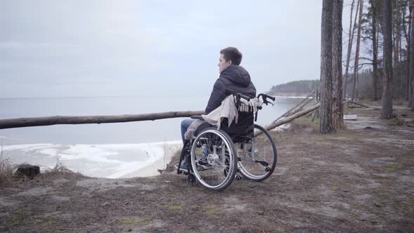 Camera Approaching To Lonely Sad Caucasian Boy in Wheelchair Looking Away. Young Crippled Man