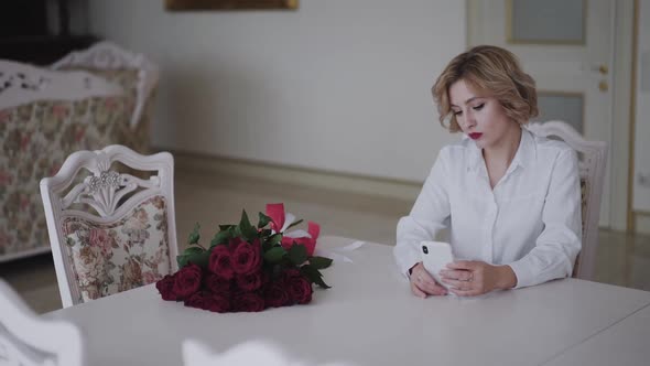 Portrait of Romantic Lady Uses Phone at Table with Bouquet of Roses
