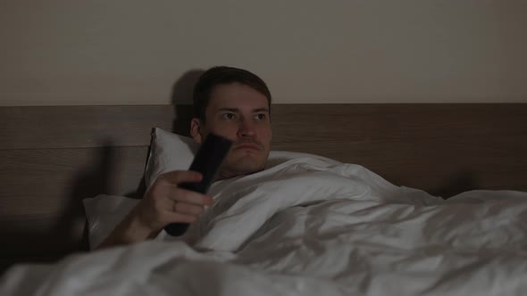 Young Man Lying on Pillow Under Blanket in Bed and Watching Tv in Late Evening