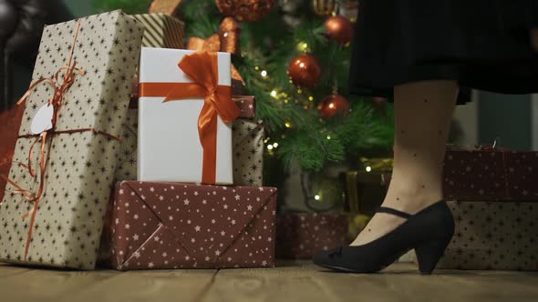 Woman Walks Up To the Christmas Tree and Takes Gift Box From Stack of Gifts Lying on the Wooden