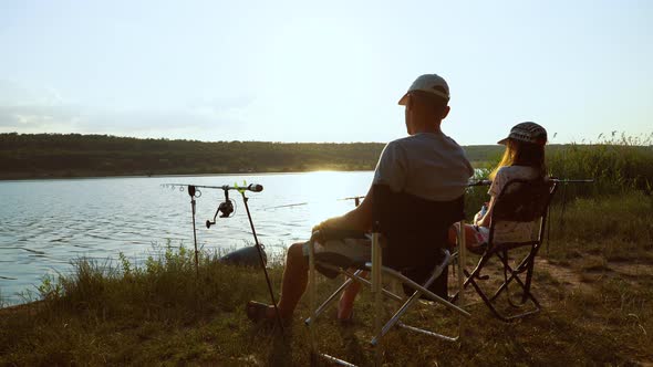 Father and daughter fishing on river bank at sunset