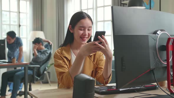 Asian Female Using Mobile Phone While Works On Her Personal Computer With Big Display In Office