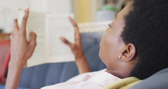 Happy african american woman lying on sofa in living room, reading book