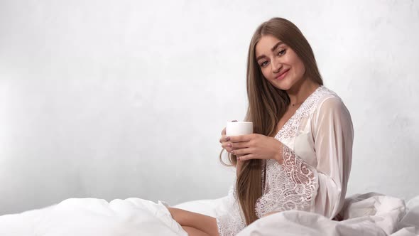 Adorable Woman in Dressing Gown Posing at White Bedroom Drink Hot Tea
