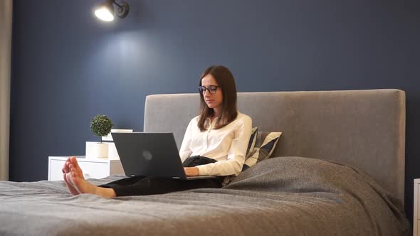 A Young Woman with Glasses Smiling While Working on a Laptop From Her Bedroom