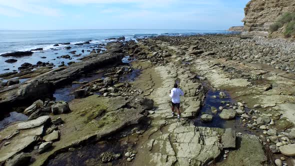 Tracking shot of a young man running on a rocky ocean beach shoreline