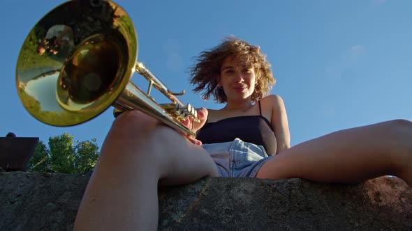 a Curly Woman Sits and Looks Into the Camera Against the Blue Sky and Holds Trumpet in Her Hand