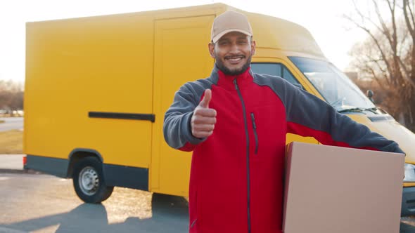 Smiling Adult Delivery Man Carrying Cardboard Box Showing Thumb Up Gesture and Looking at Camera