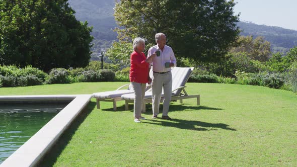 Happy caucasian senior couple walking in sunny garden holding hands carrying cups and talking