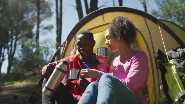 Smiling diverse couple sitting in tent and drinking tea in countryside