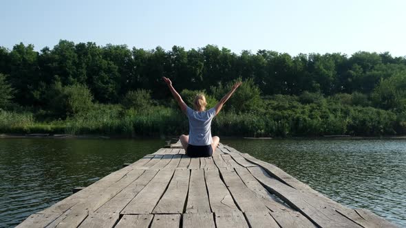 Young Woman Sitting on a Wooden Bridge Over the River and Practices Yoga in the Early Morning