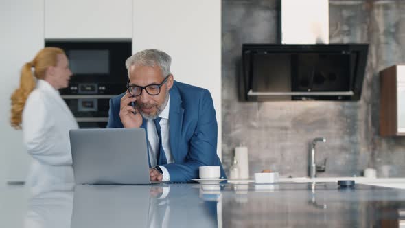 Confident Mature Entrepreneur Working on Laptop and Talking on Phone in Kitchen in Morning