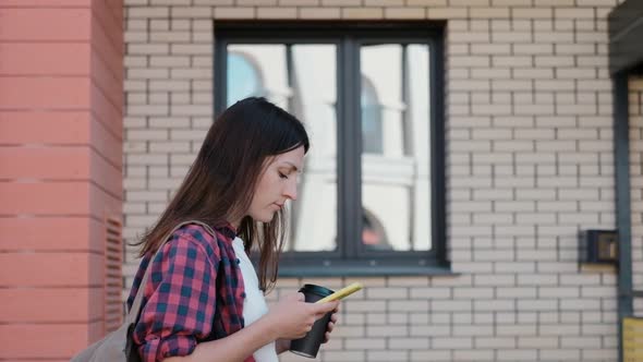 Smiling Young Woman Wearing Glasses Uses Smartphone Looking at the Screen While Walking Around the