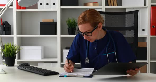 Doctor in Blue Lab Coat Sitting at Workplace and Marking Documents
