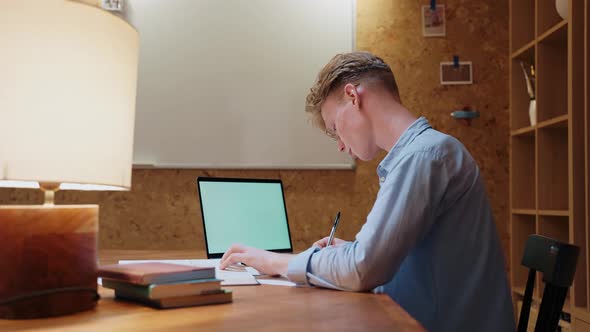 Young Man Studying at Home Writes in a Notebook and Reads a Textbook