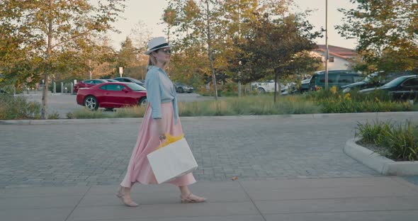 Woman with Bags Walking By Street Parking Lot at Shopping Mall on Summer Evening