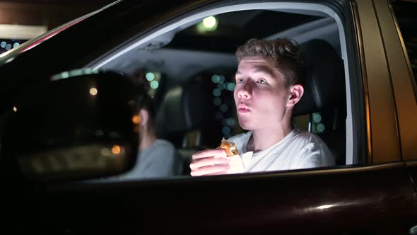 Young guy eating burger in the car driving late at night in the parking lot