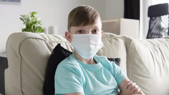 A Young Boy in a Face Mask Looks at the Camera As He Sits on a Couch at Home - Closeup