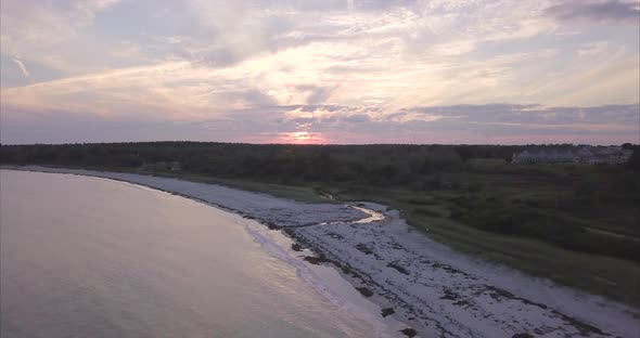 Aerial shot over a small stream flowing out to sea on the Maine coast with a cloudy orange sunset in