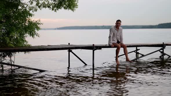 Young Man is Relaxing on Wooden Pier in Early Morning Rest in Nature in Summer