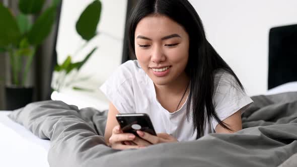 Asian Lovely Brunette Girl Lies on a Cozy Bed at Home Lies on Her Stomach Uses Smartphone Texting