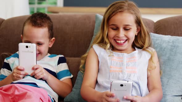 Smiling boy and girl sitting on sofa and using mobile phone