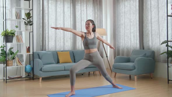 Asian Athletic Female Exercising, Stretching And Practicing Yoga In Warrior Ii Pose At Home