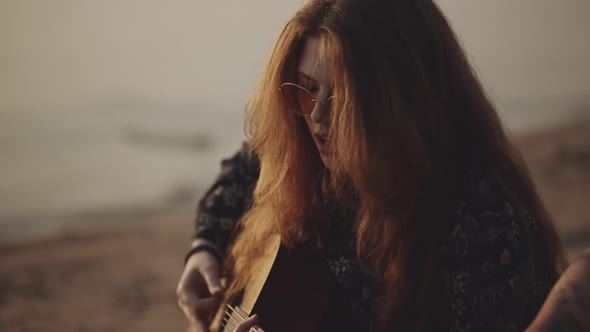 Red-haired Hipster Girl in Beautiful Sunglasses Plays the Guitar. Pretty Female Sings a Song on