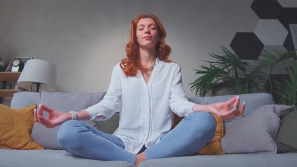 Caucasian Woman Doing Yoga Sits on Sofa in Lotus Position in Living Room