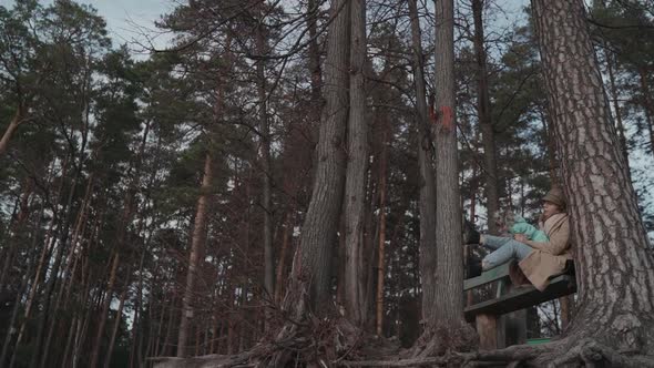 Young Woman in Coat is Sitting on Bench in Pine Forest with Dog on Her Lap Enjoying Nature