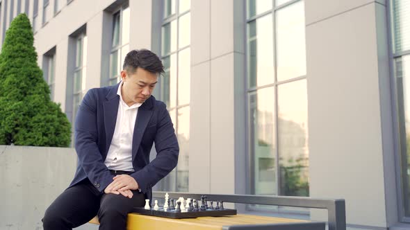 Concentrated Young Asian businessman playing chess on a bench on an urban