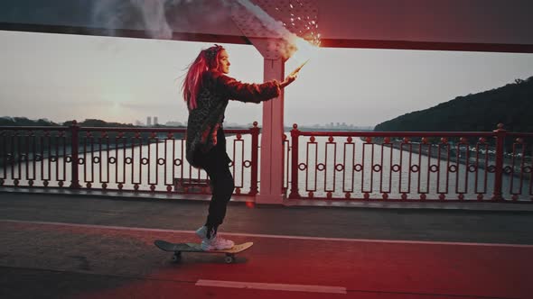 Young Hipster Female in Informal Outfit is Riding Skateboard on Bridge Holding Burning Red Signal