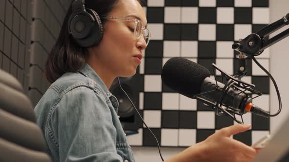 Confident young Asian woman talking into microphone while recording radio show and texting by phone