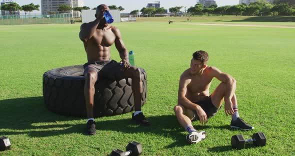 Two fit, shirtless diverse men resting, drinking water and bumping fists after exercising outdoors